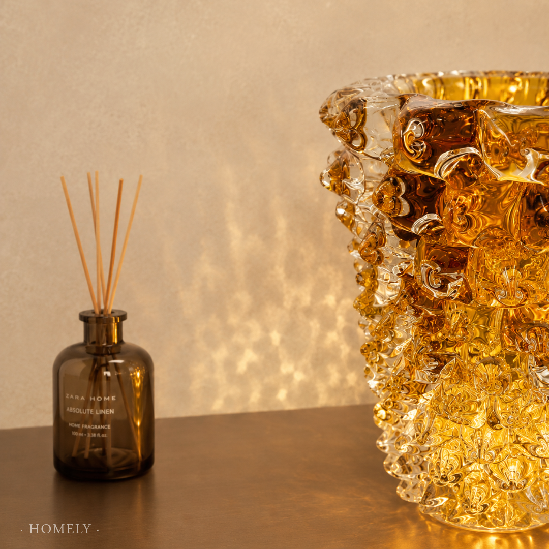 Brown diffuser bottle with sticks and decorative glass vase on a reflective surface with a beige background