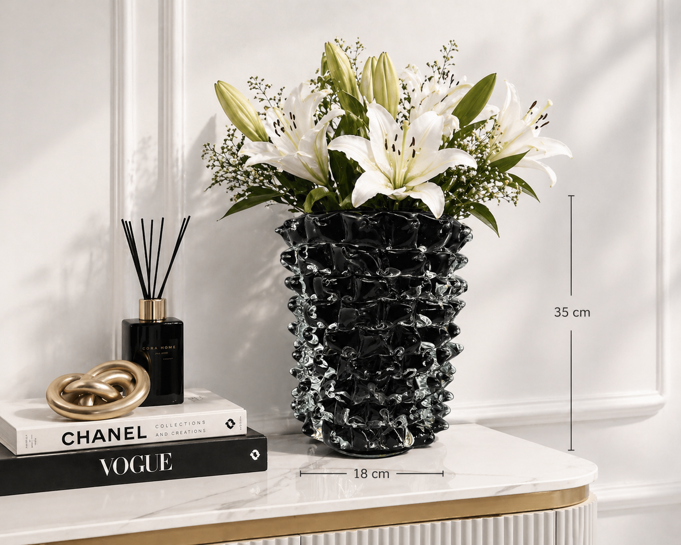 Black textured vase with white flowers on a shelf with books and a diffuser.